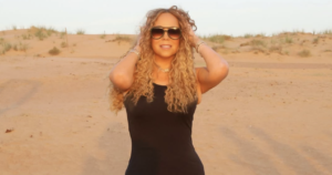 Mariah Carey posing in a desert at sunset, wearing a short black dress, oversized sunglasses, and unique, waist-high black boots.