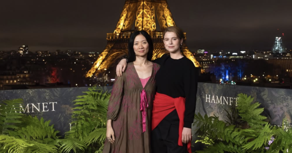 Two women stand arm-in-arm on a green terrace at night, with the illuminated Eiffel Tower glowing brightly behind them. Lush ferns surround them, and the word "HAMNET" is visible on the backdrop.