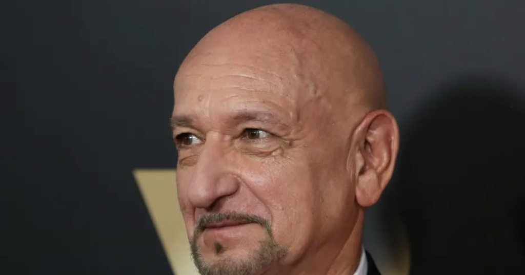 A close-up portrait of Sir Ben Kingsley, featuring his signature bald head and a neatly trimmed salt-and-pepper goatee, wearing a black suit with a white shirt and maroon tie.