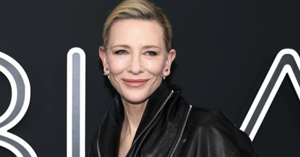 Cate Blanchett smiling at a red carpet event, wearing a structured black leather jacket with a high collar and cinched waist against a black background with white lettering.
