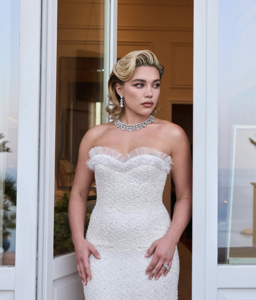 Actor Florence Pugh standing in a white doorway wearing a strapless, heavily embellished white lace gown with a ruffled sweetheart neckline, paired with a sparkling diamond statement necklace and matching drop earrings, looking off to the side.