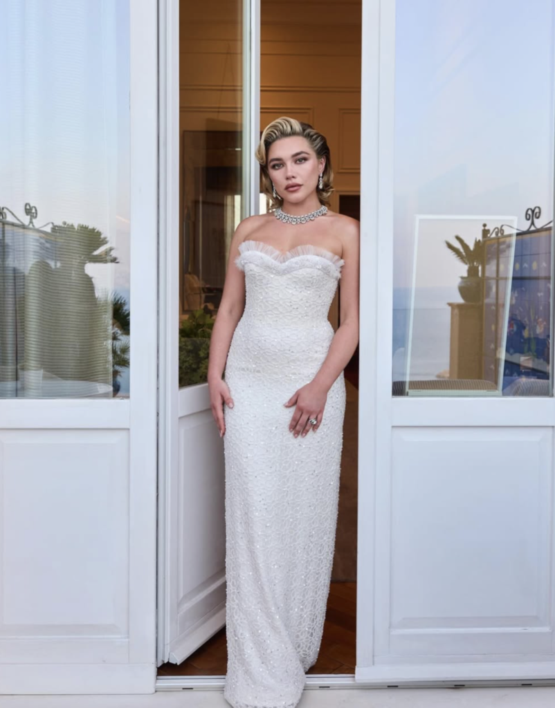 Full-length view of actor Florence Pugh standing between open white doors, wearing a floor-length, form-fitting white lace and sequin strapless dress. She has styled blonde hair and is wearing a prominent diamond necklace, looking directly at the camera.