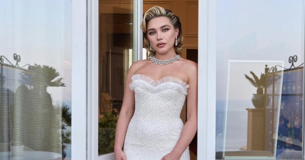 Full-length view of actor Florence Pugh standing between open white doors, wearing a floor-length, form-fitting white lace and sequin strapless dress. She has styled blonde hair and is wearing a prominent diamond necklace, looking directly at the camera.